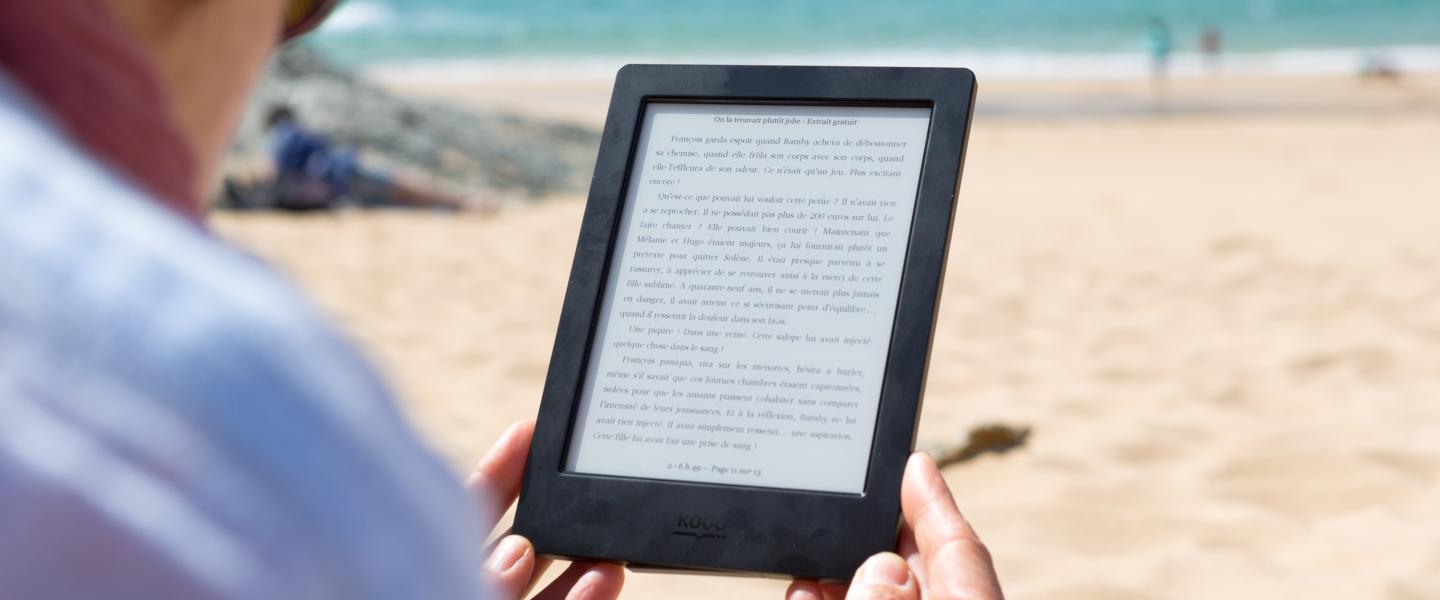 Kuva naisesta rannalla lukemassa e-kirjaa tabletilla