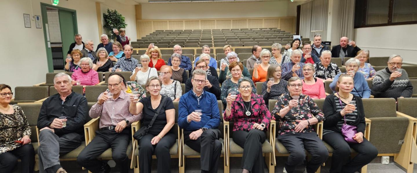 Auditoriossa istuu ryhmä ihmisiä kuohuviinilasit käsissään