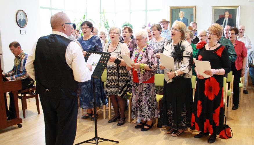 Laulavia eläkeläisiä, joita pianisti säestää ja mies johtaa