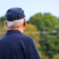 Harmaahiuksinen mies lippalakki päässä katsoo eteenpäin. Kuvattu viistosti takaa. Taustalla puita.
