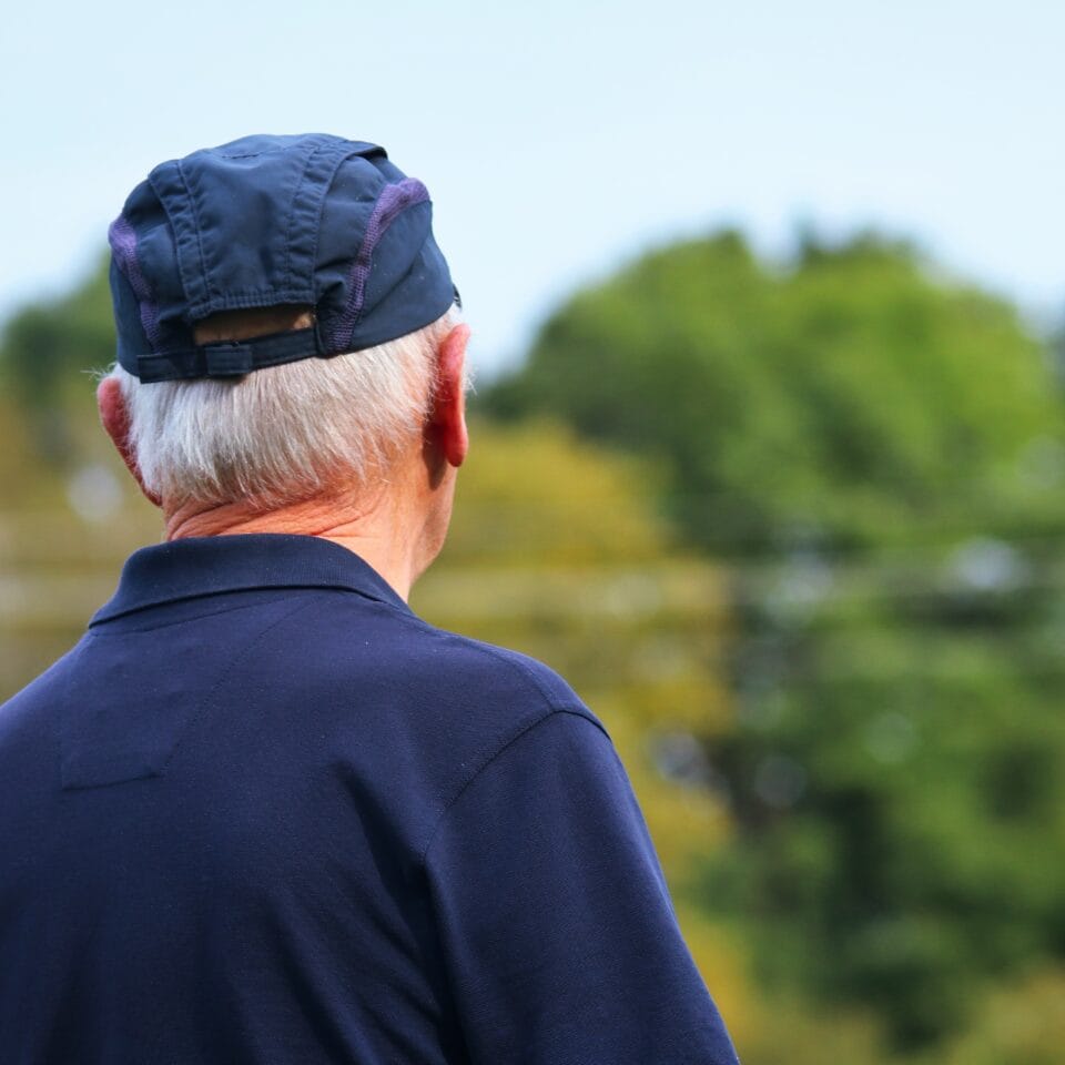 Harmaahiuksinen mies lippalakki päässä katsoo eteenpäin. Kuvattu viistosti takaa. Taustalla puita.