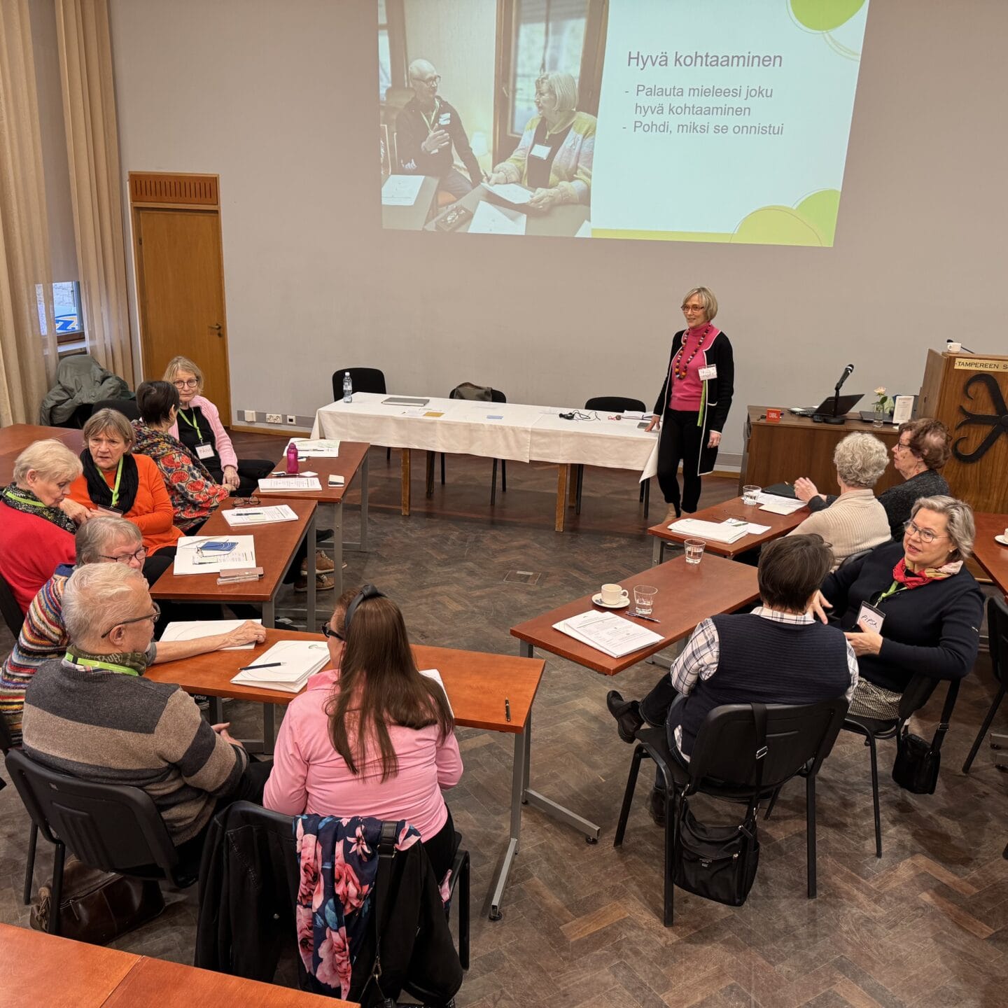 Vapaaehtoiset istuvat u-muodostelmassa pöytien vieressä ja pohtivat pareittain Lähettiläsvalmennuksessa saamaansa tehtävää. Kuva on otettu viistosti ylhäältä ja siinä näkyy koko n.10 henkinen ryhmä sekä taka-alalla yksi valmentaja sekä seinälle heijastettu tehtävädia.
