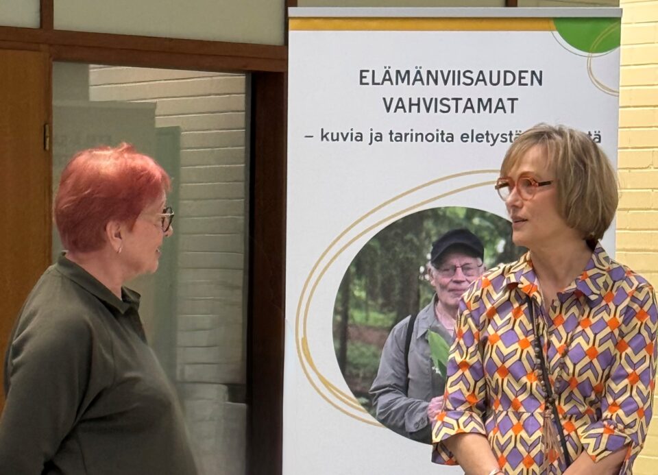 Ylävartalokuva vapaaehtoisesta Maire Vierikosta, jota projektisuunnittelija Teija Saarisen haastattelee. Taustalla Elämänviisauden vahvistamat -näyttelyn roll up.